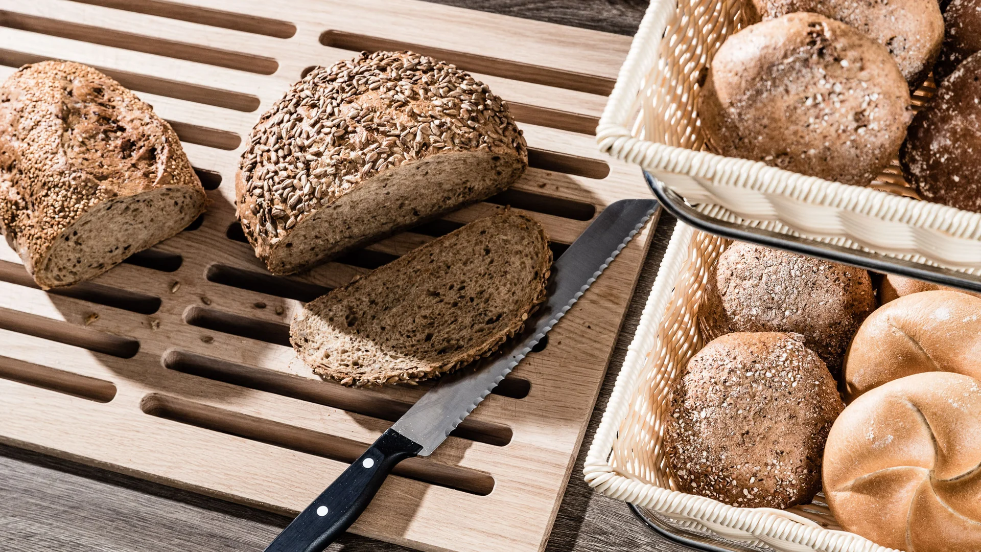 Galleria: impressioni dal Plattenhof a Termeno Varie tipologie di pane su un tagliere di legno e in un cestino, con coltello da pane.