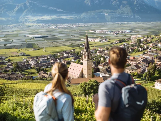 Zwei Wanderer blicken auf Dorf mit Kirche und Berge im Hintergrund