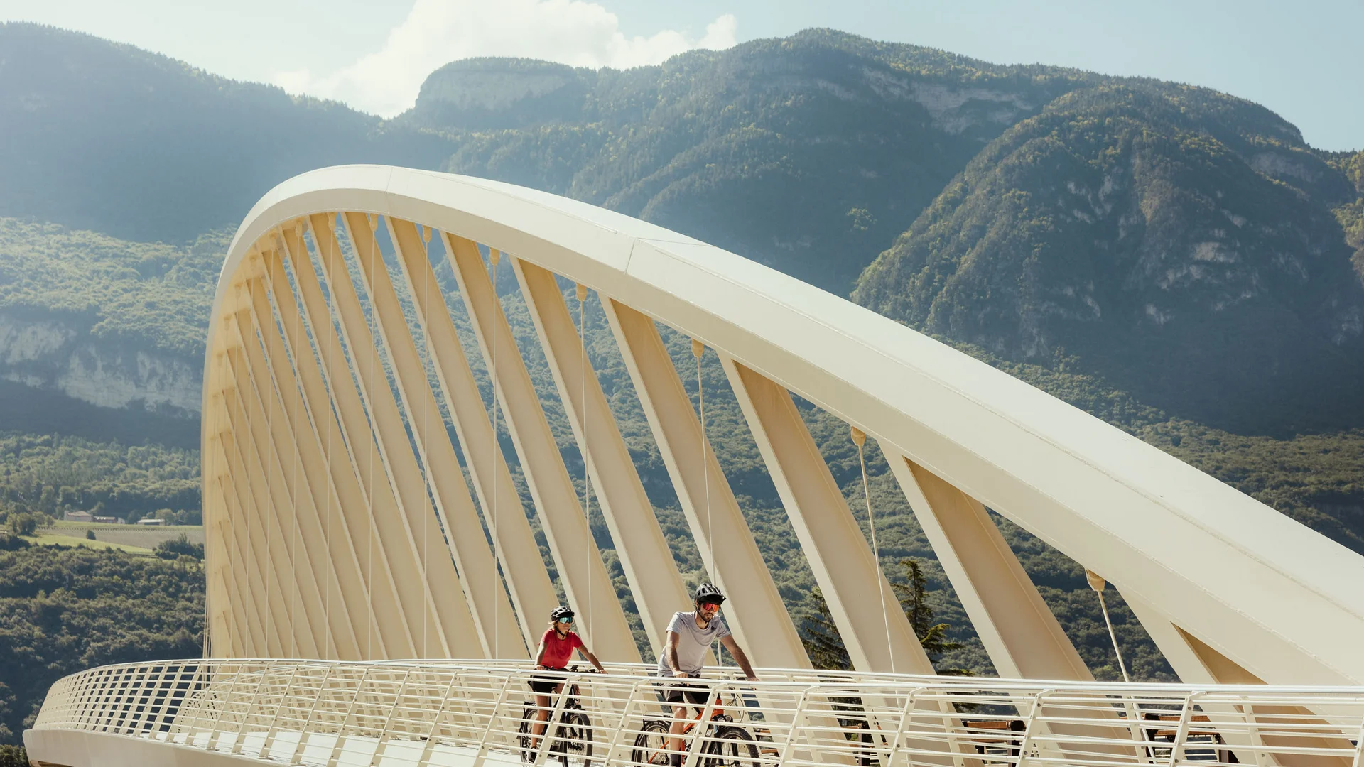 Galleria: impressioni dal Plattenhof a Termeno Due ciclisti su un ponte moderno davanti a un paesaggio montano